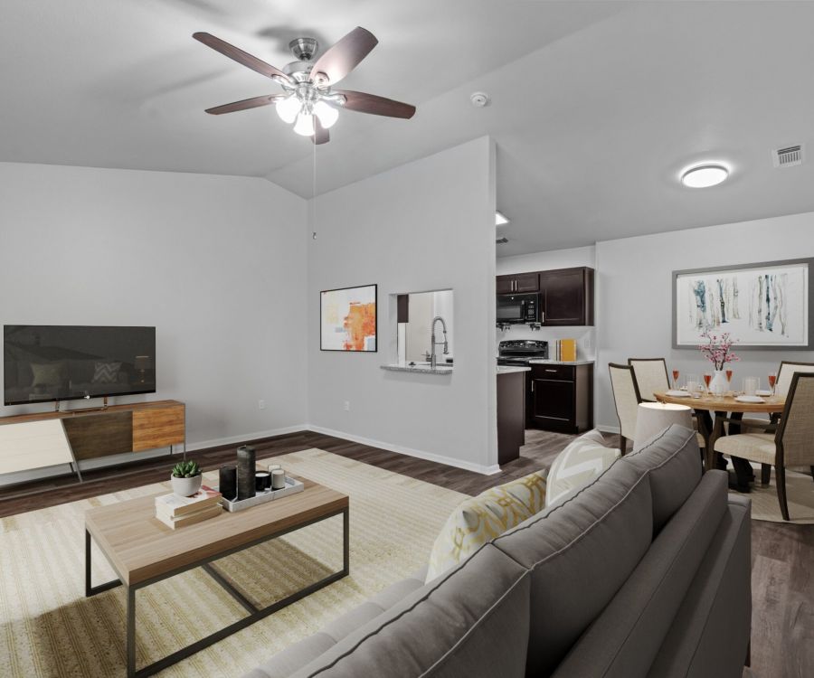 Modern living room in Encino Park apartments with gray couch, TV, and dining area. Ceiling fan above, kitchen in background. Bright decor.