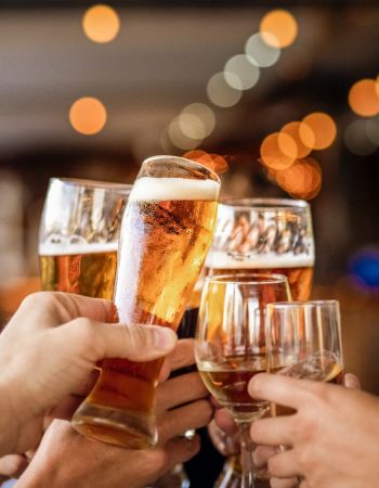 People clinking glasses filled with beer and wine in a celebratory toast, with blurred lights in the background.
