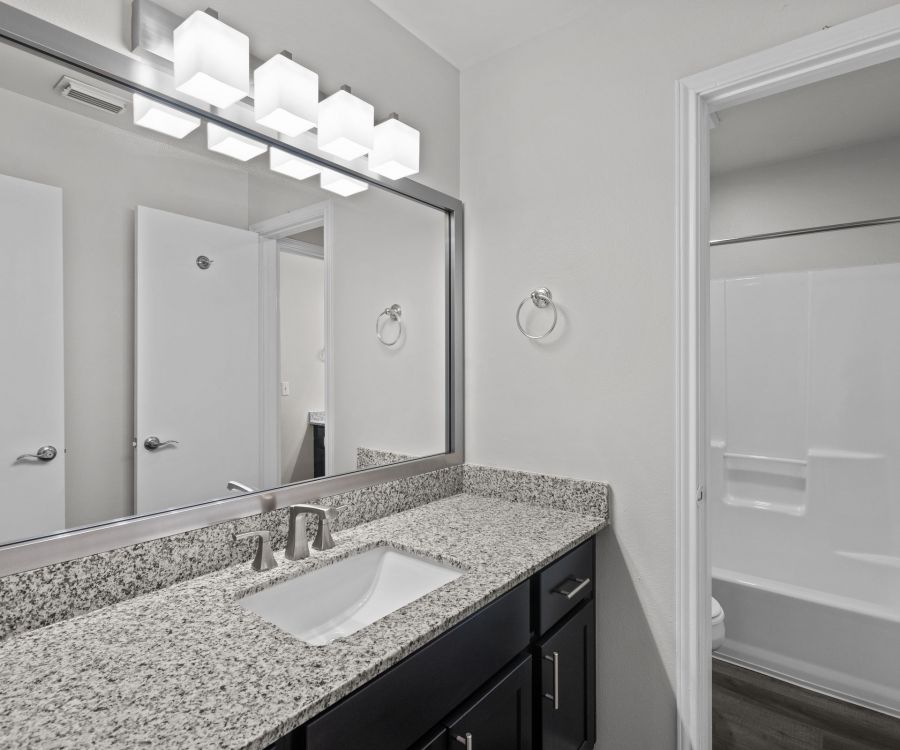 Modern bathroom in Encino Park Apartments with a granite countertop, large mirror, and bathtub. Bright lighting fixtures above the mirror.