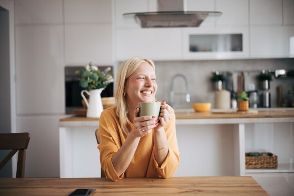 A woman in a yellow sweater smiles while holding a mug in a modern kitchen.