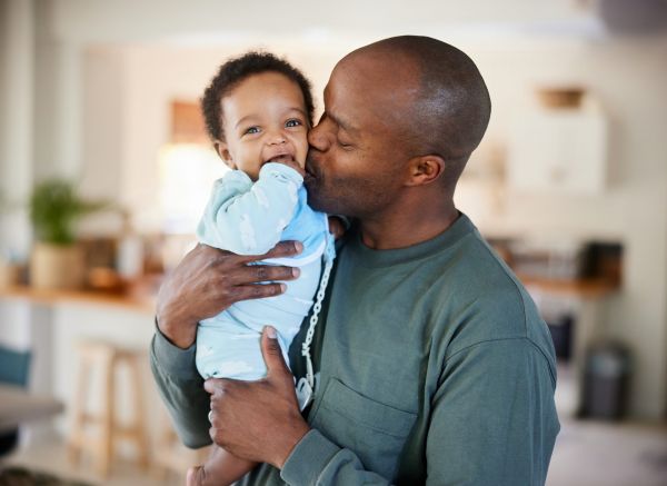 A man lovingly kisses a smiling baby in a home setting, both dressed in comfortable clothing.